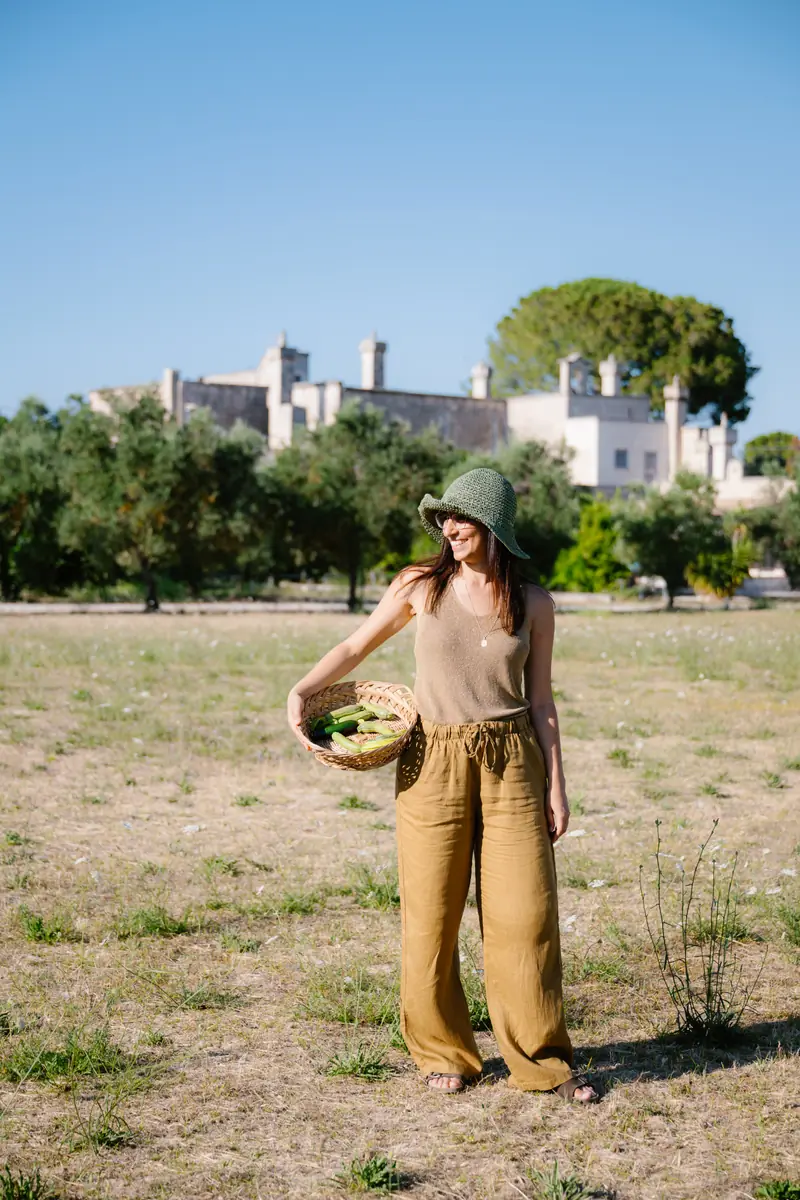 Simona con un cappello di paglia e un cesto di zucchine in un campo, con un edificio storico sullo sfondo